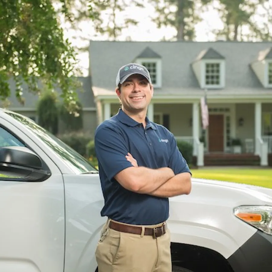 Home service technician in uniform standing with arms crossed by a company vehicle in front of a suburban house.