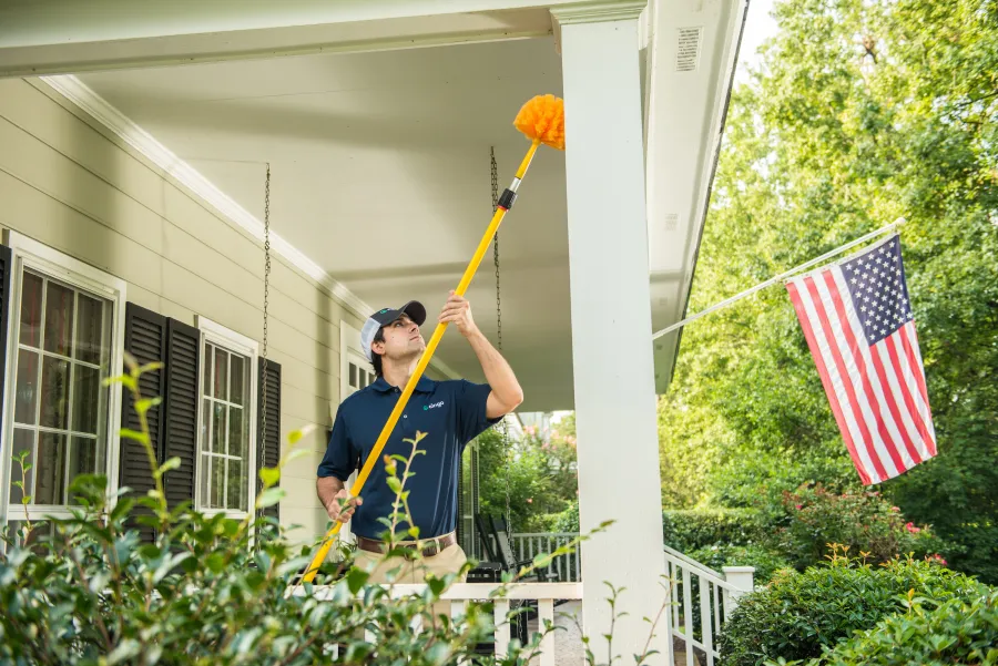 Man cleaning porch ceiling with a long yellow duster beside an American flag on a sunny day.