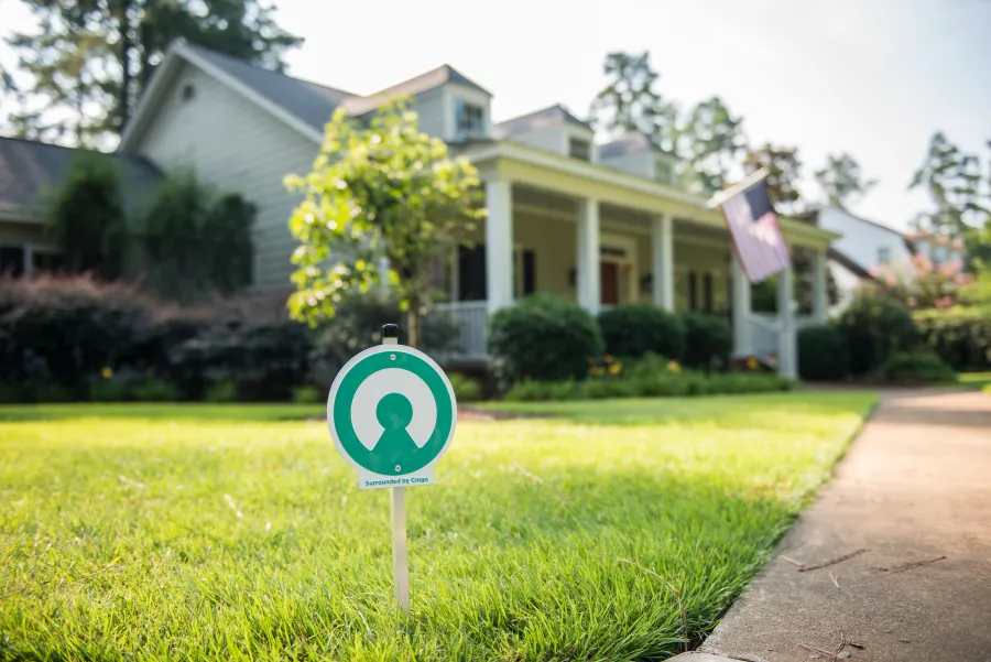 Security system sign planted on a green lawn in front of a suburban house with a porch and American flag.