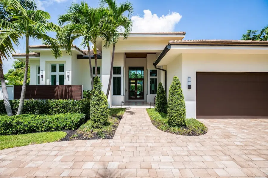 Modern white house with large windows, palm trees, and a paved driveway under a blue sky