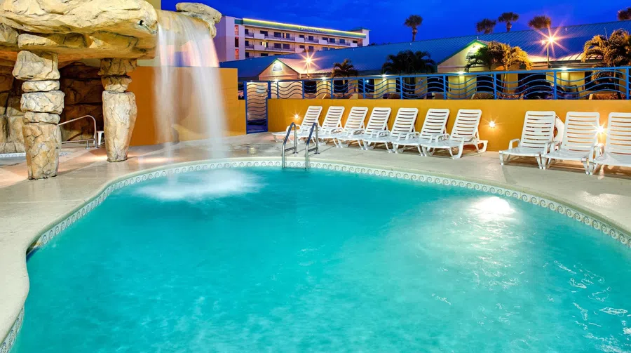 Outdoor hotel pool at dusk with waterfall feature and white lounge chairs under bright lights.