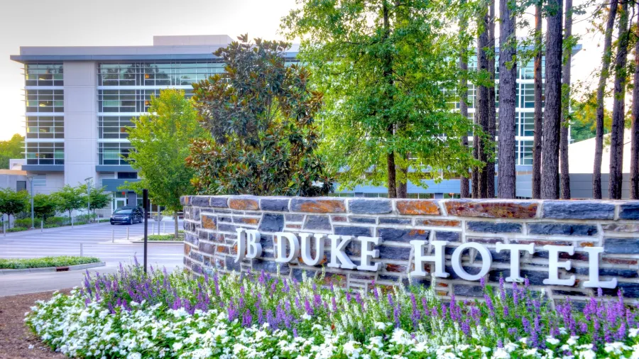 Stone sign for JB Duke Hotel surrounded by purple and white flowers with modern building and trees in the background