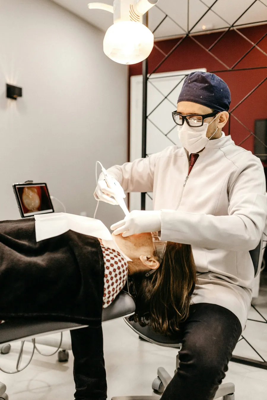 Dentist wearing mask and gloves treating a patient lying on dental chair with equipment and screen in clinic.
