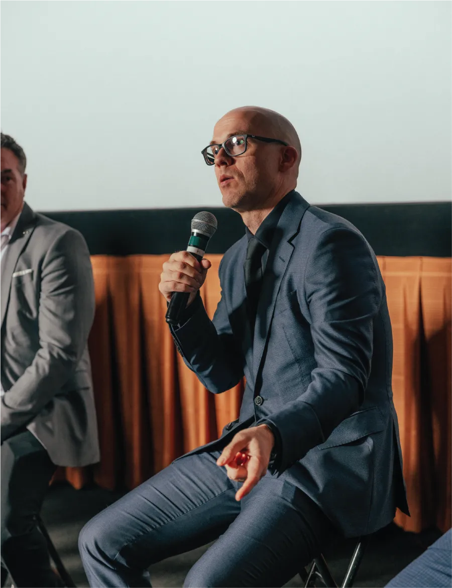Bald man in glasses speaking into a microphone during a panel discussion indoors, dressed in blue suit and black tie.