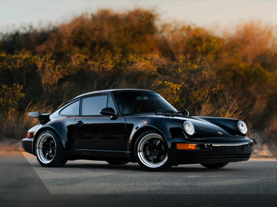 Black classic Porsche 911 sports car parked on pavement with autumn trees in the background at sunset.