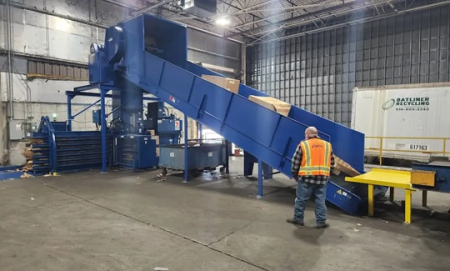 Worker in safety vest operates blue cardboard baler conveyor inside industrial recycling facility.