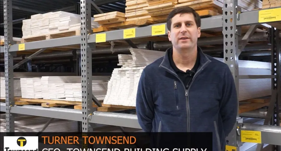 Man stands in warehouse with wood shelves behind him, visible video editing software on computer screen beside him.