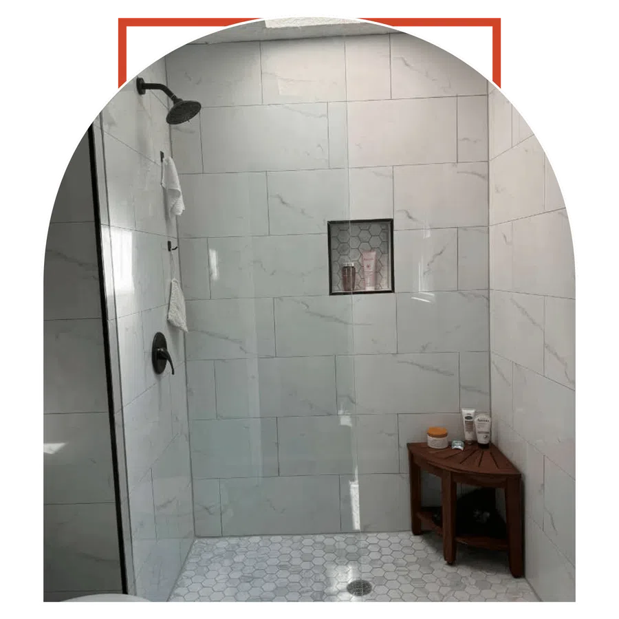 Modern shower with white marble tiles, hexagon floor tiles, black fixtures, wooden corner bench, and toiletries.