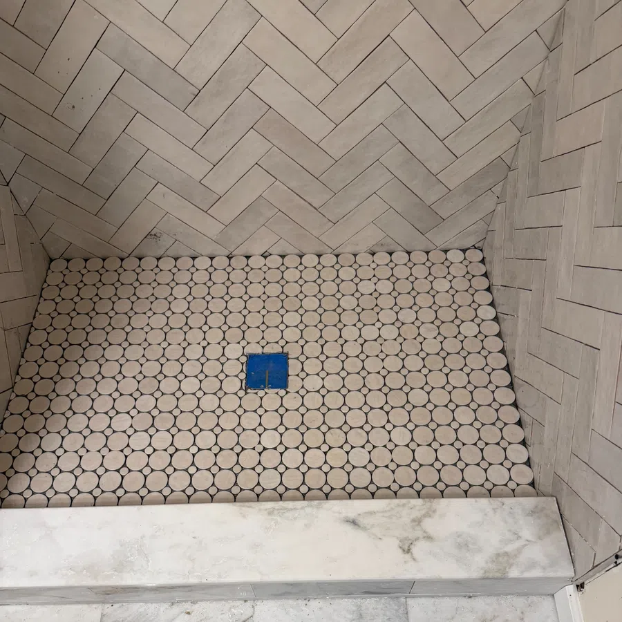 Shower floor with round white tiles and blue drain surrounded by beige herringbone patterned wall tiles.