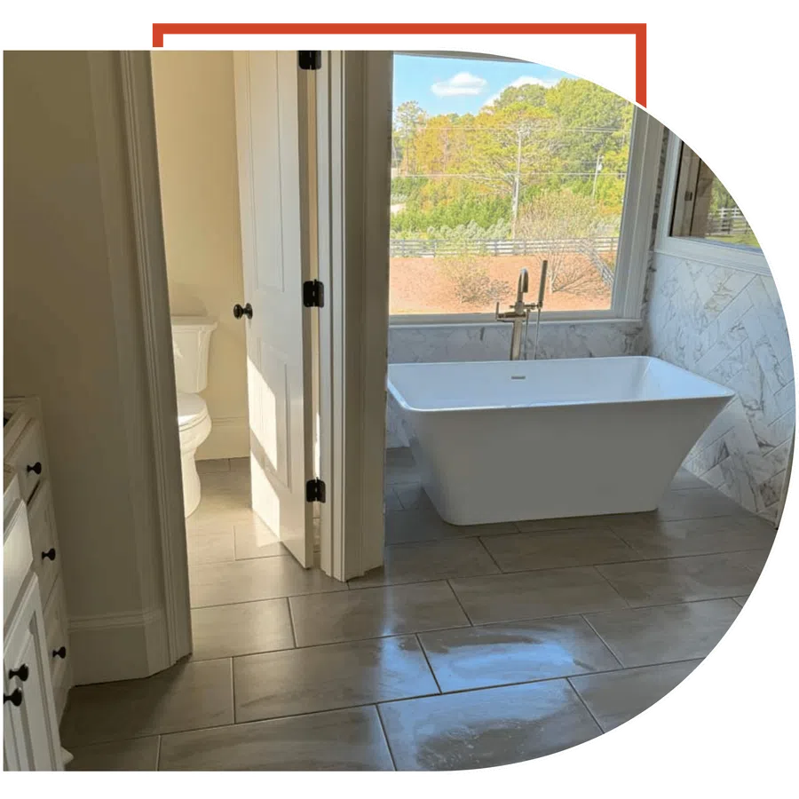 Modern bathroom with freestanding tub, large window, marble wall tiles, and adjacent toilet room with door.