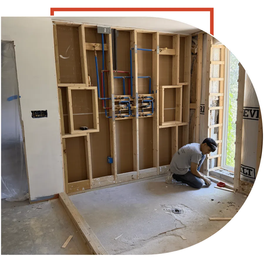 Worker installing plumbing pipes inside wooden frame wall during home renovation near large window.