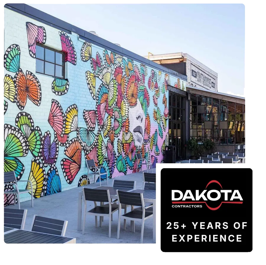 Outdoor cafe seating next to a colorful mural of a face surrounded by vibrant butterfly wings on a building wall.