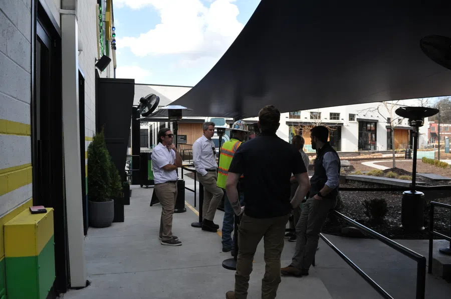 Group of people, including one in a safety vest, talking under a large black canopy outside modern buildings on a sunny day