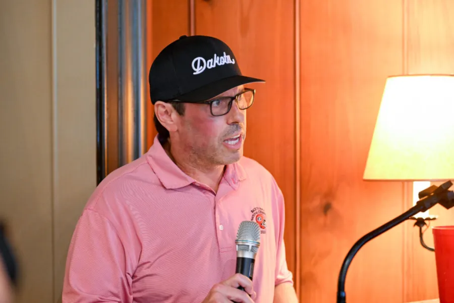 Man in pink polo and black cap speaking into handheld microphone indoors near lamp and wooden wall