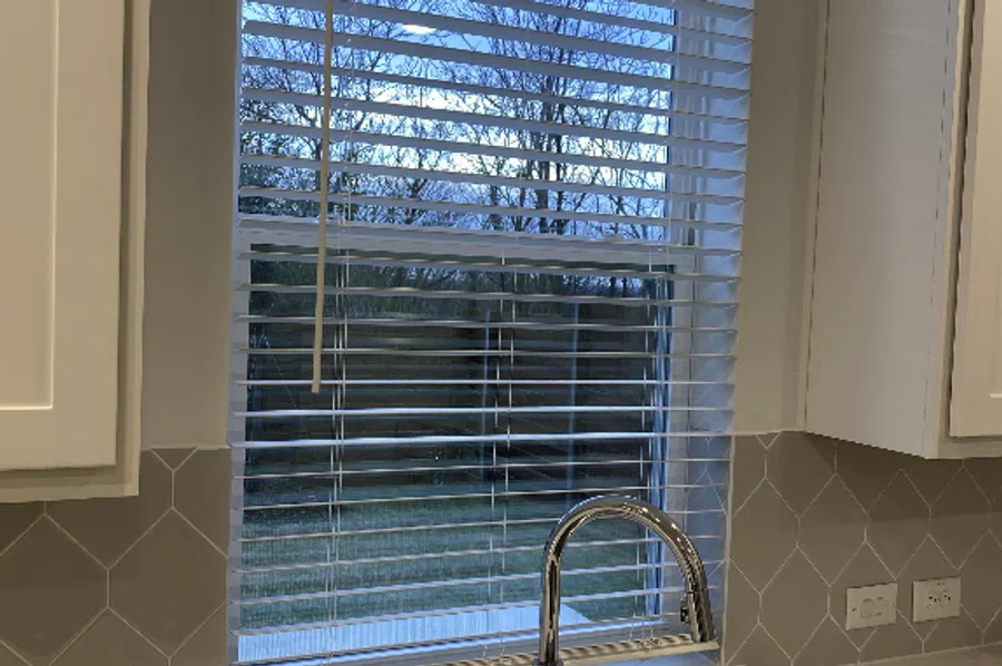 Kitchen window with white blinds overlooking a backyard, hexagonal tile backsplash, and stainless steel faucet