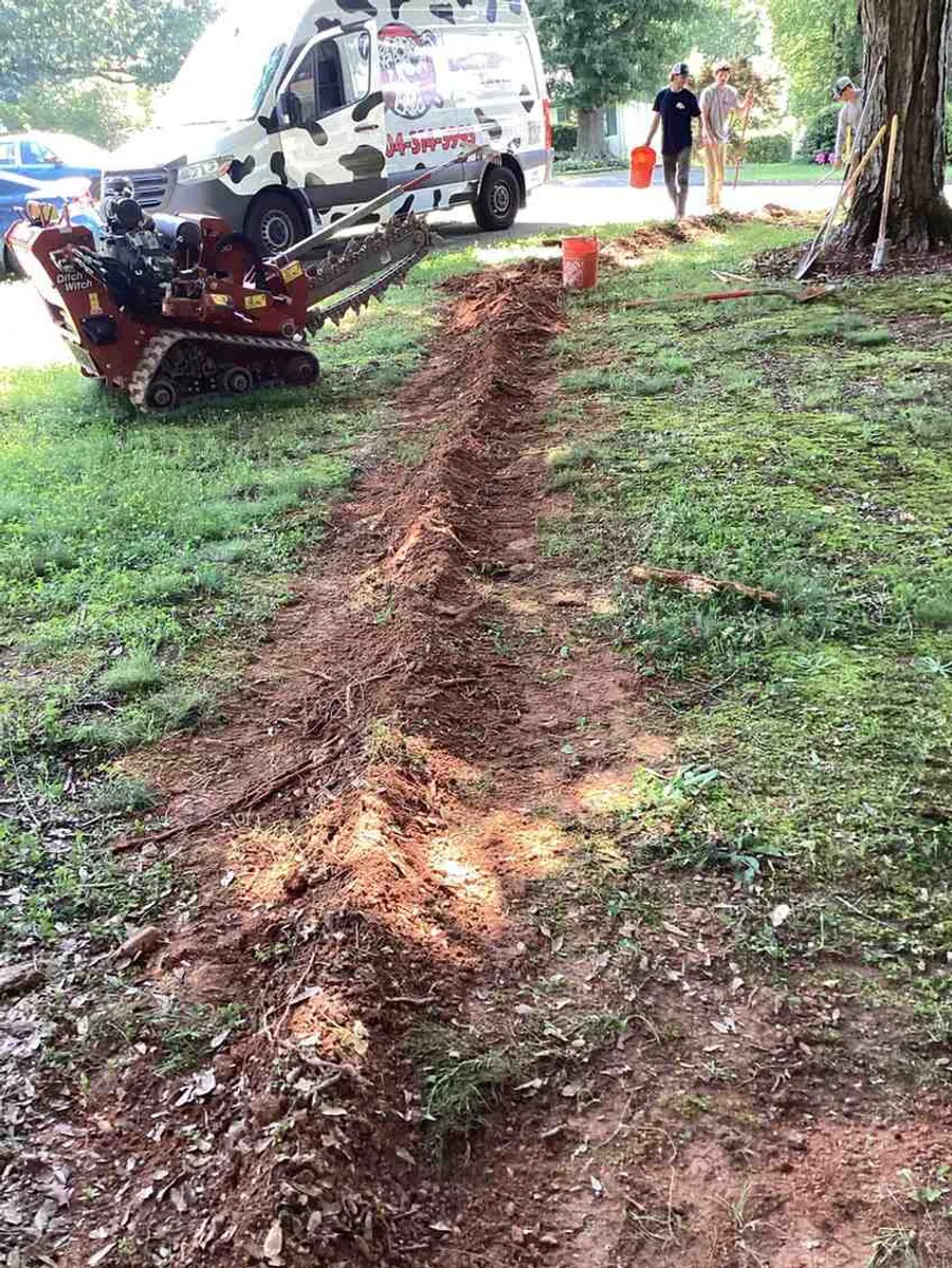Trench dug in grass with mini trencher machine nearby and two workers carrying orange buckets in background.