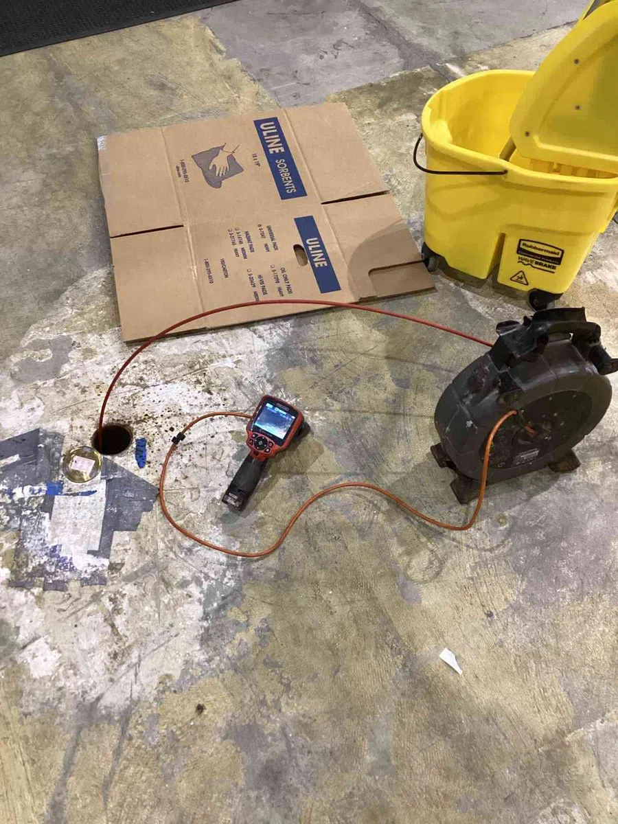 Inspection camera with cable, cardboard, and yellow mop bucket on worn concrete floor in industrial setting