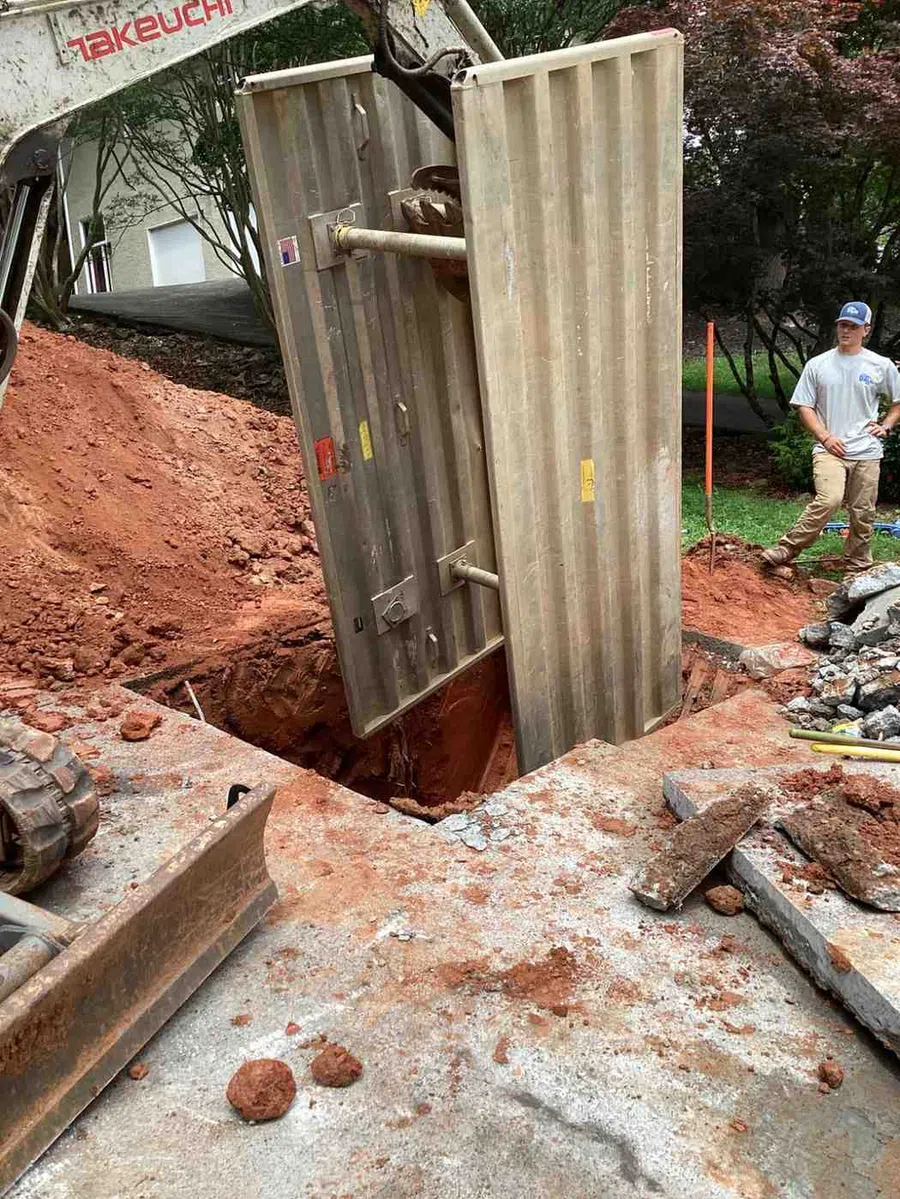 Excavator lowering a large trench box into a deep rectangular excavation site with a worker standing nearby.