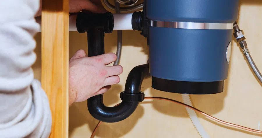 Hand fixing black pipes under kitchen sink connected to a blue garbage disposal unit inside wooden cabinet