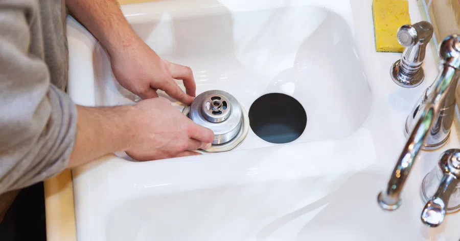 Person installing or repairing a metal drain stopper in a white kitchen sink with chrome faucet and yellow sponge.