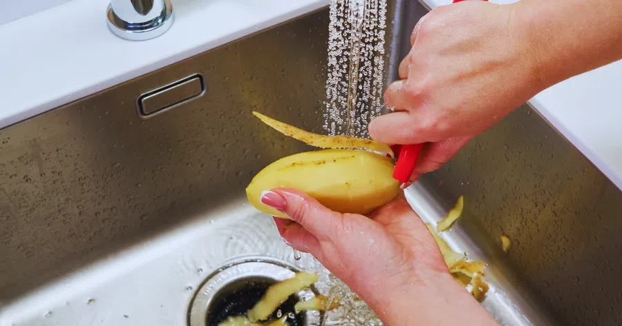 Person peeling a yellow potato under running water in a stainless steel kitchen sink.