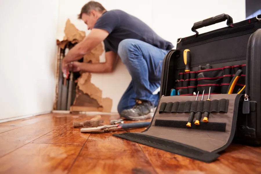 Handyman repairing damaged wall behind wooden floor with tools and toolbox in foreground