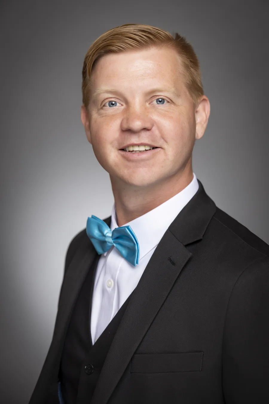 Smiling man with Red hair wearing a black suit, white shirt, and blue bow tie with a neutral gray background