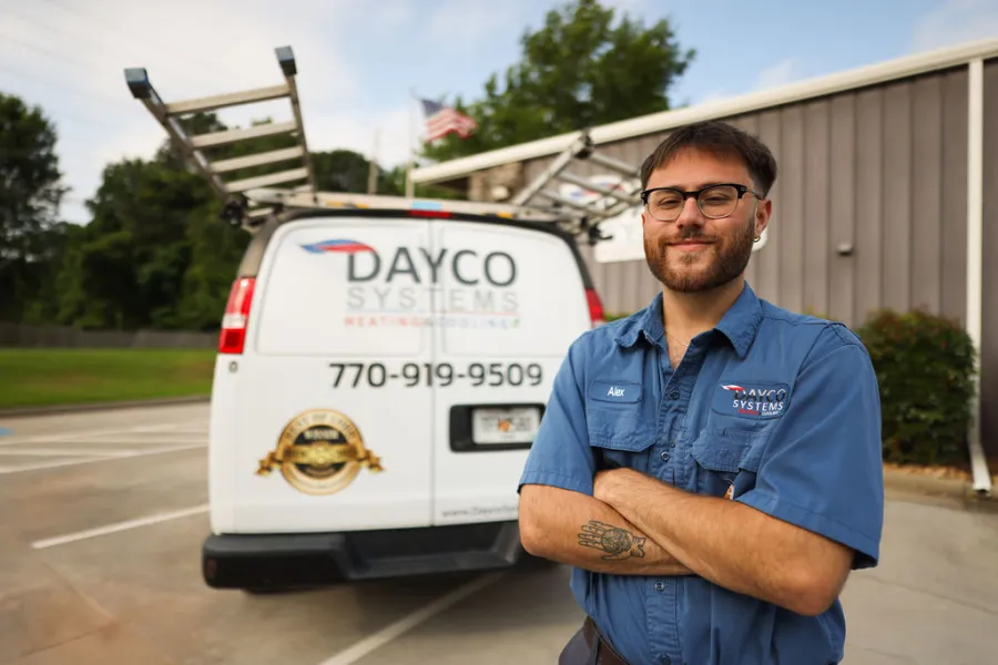 Dayco Systems technician with folded arms stands in front of a branded service van with ladders and phone number.