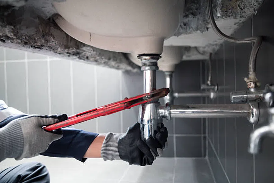 Plumber using a red pipe wrench to repair chrome pipes under a sink in a tiled bathroom area