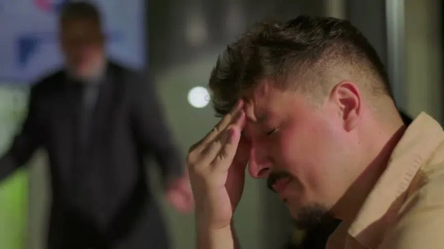 Stressed man holding his forehead with eyes closed while another person blurred in background.