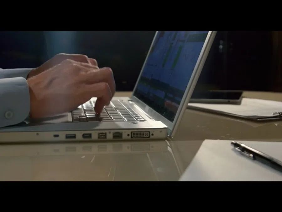 Person typing on a laptop keyboard in an office setting with paperwork and pen nearby.