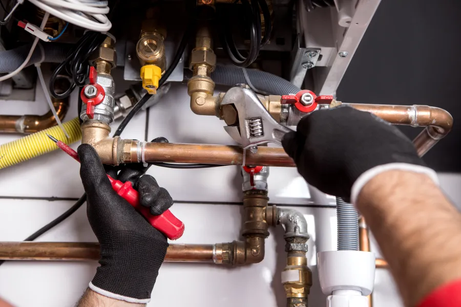 Plumber wearing black gloves fixing copper pipes using wrench and testing tool in boiler system.