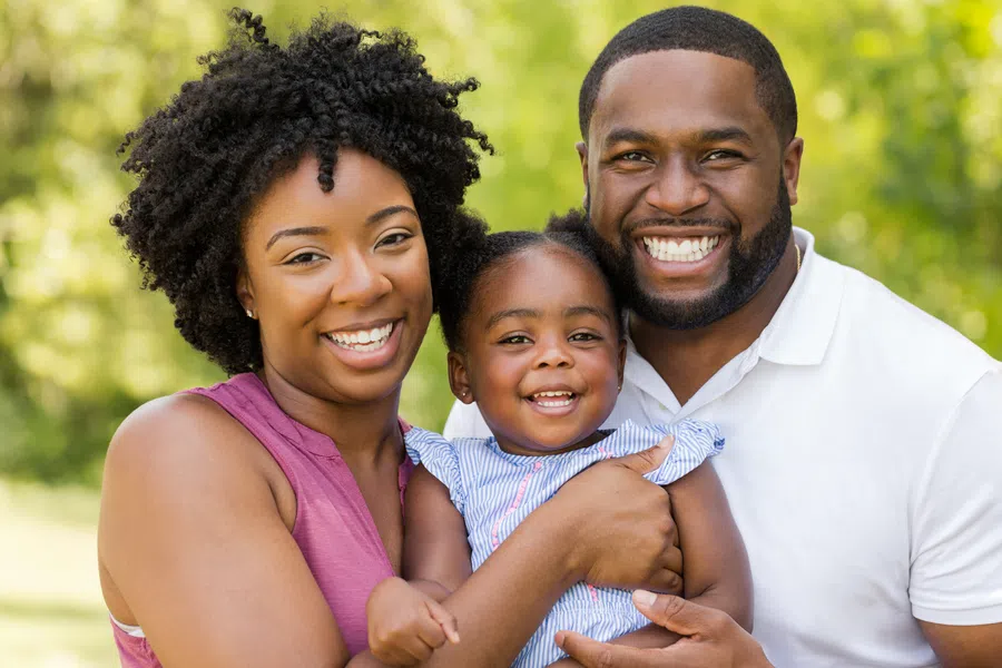 Happy African American family of three smiling outdoors with green blurred background, embracing and enjoying time together