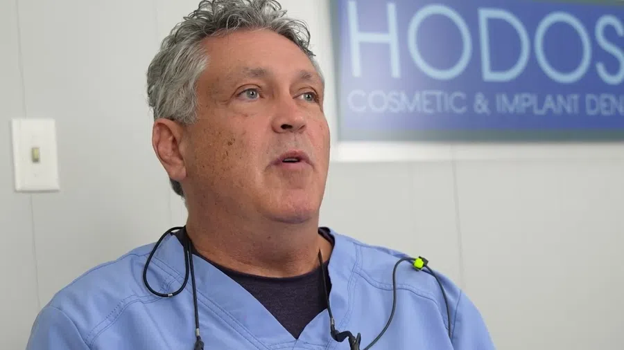 Middle-aged male dentist in blue scrubs speaking with dental clinic sign in background