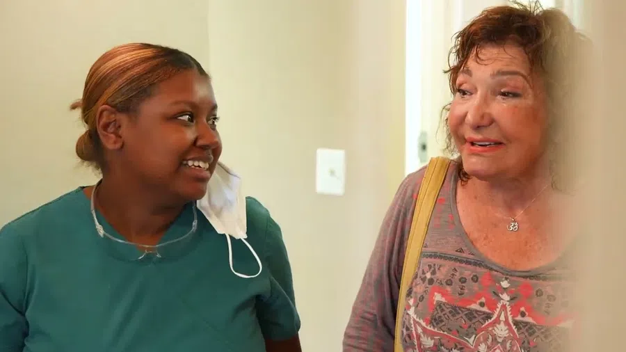 Healthcare worker in scrubs smiling and talking with an elderly woman in a casual setting indoors.