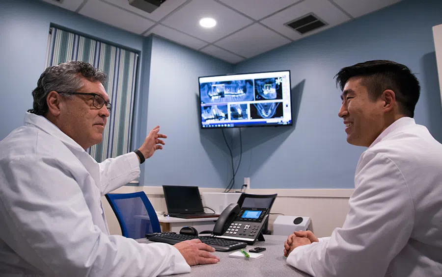 Two male doctors in white coats discussing dental X-rays displayed on a wall monitor in a medical office.