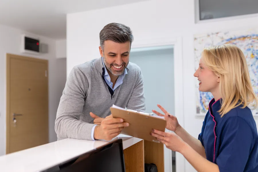 Smiling man consulting with receptionist holding clipboard at a modern clinic reception desk.