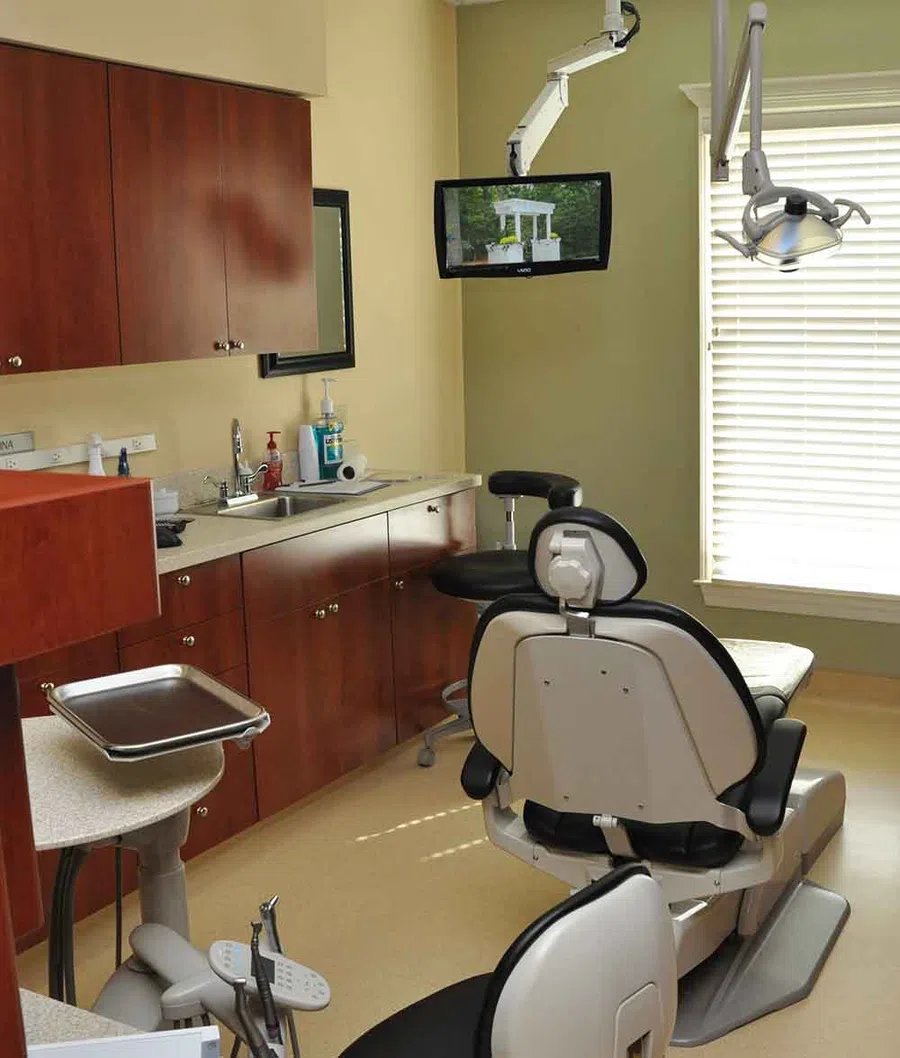 Modern dental clinic room with dental chair, overhead light, TV, and wooden cabinetry under bright window light