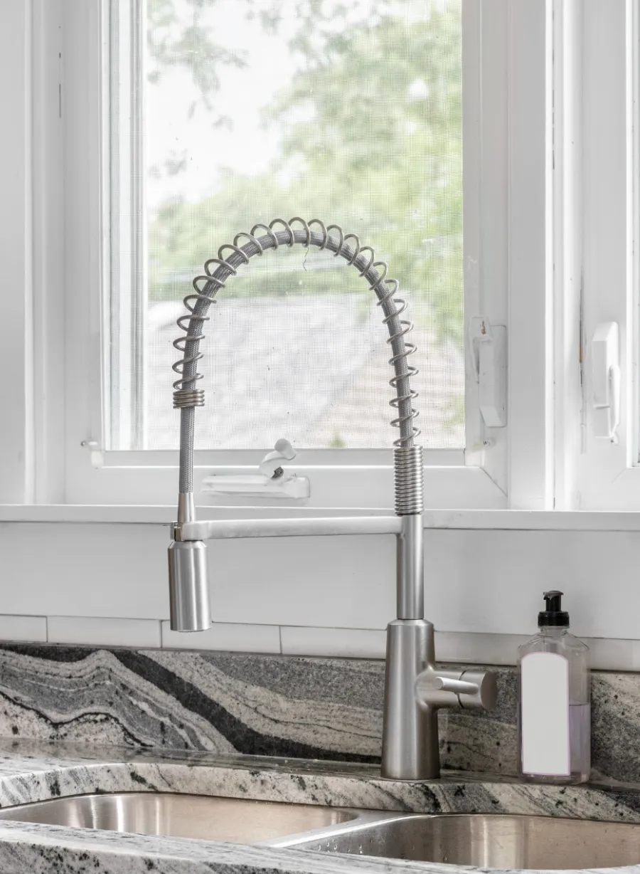 Modern kitchen sink with stainless steel faucet and granite countertop under bright windows with white tile backsplash.