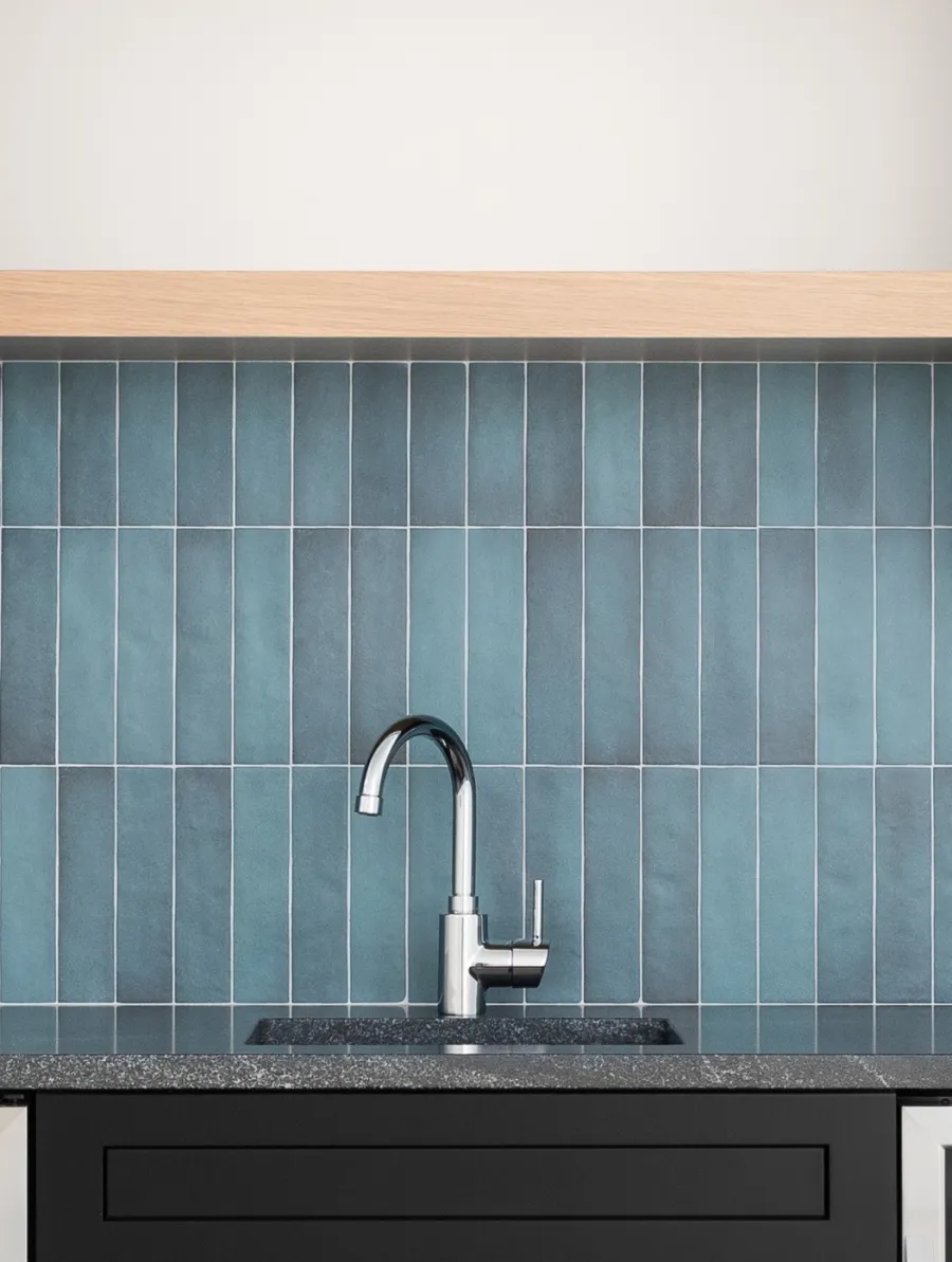 Modern kitchen sink with black cabinets, speckled countertop, and blue vertical tile backsplash under wooden shelf.