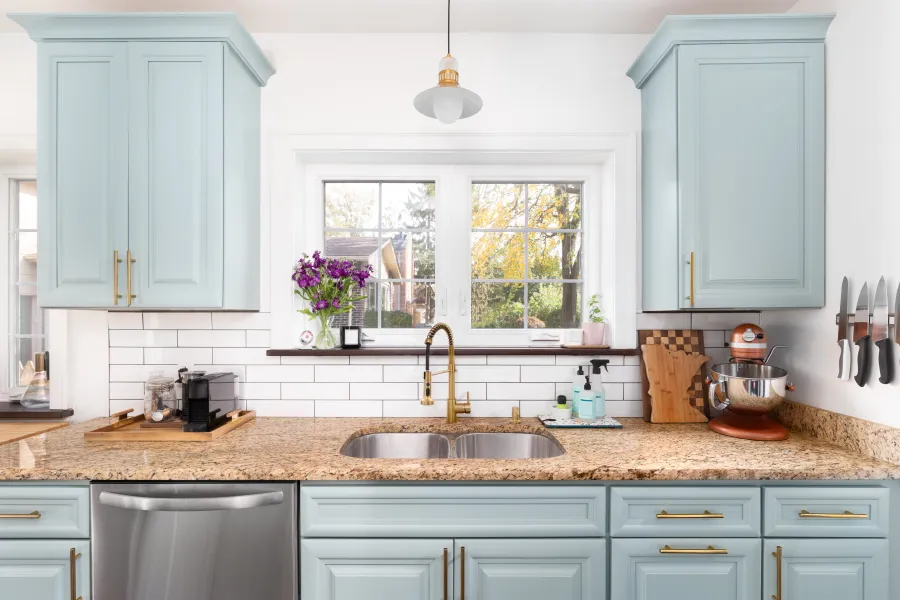 Bright kitchen with light blue cabinets, granite countertops, double sink, and stainless steel dishwasher under window.