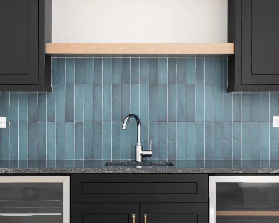 Modern kitchen sink with black cabinets, speckled countertop, and blue vertical tile backsplash under wooden shelf.