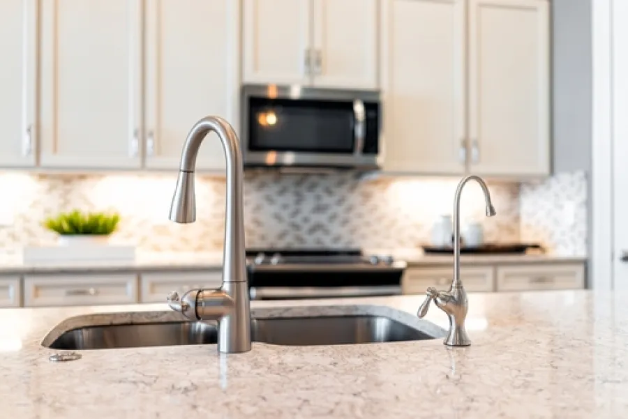 Modern kitchen sink with stainless steel faucets on marble countertop and white cabinetry in background.