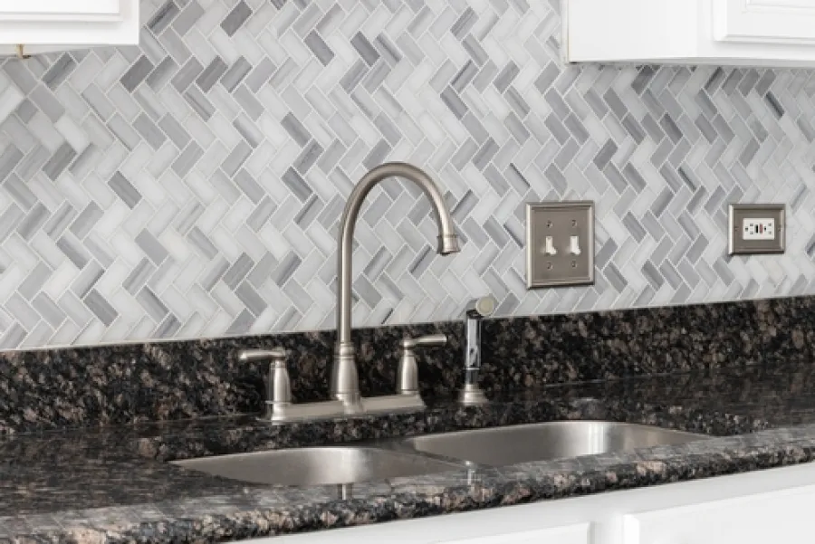 Modern kitchen sink with brushed nickel faucet and herringbone gray marble backsplash over dark granite countertop.