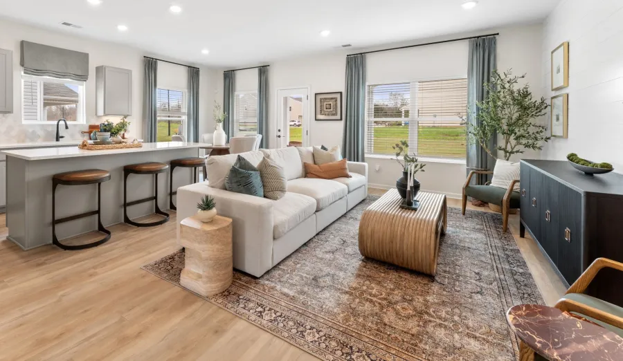 Bright open living room and kitchen with beige sofa, wooden bar stools, large windows, and stylish decor.