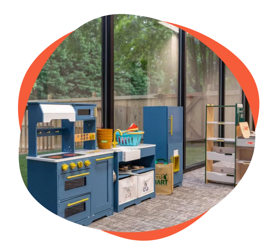 Blue toy kitchen set with stove, sink, and refrigerator in a sunlit room with large windows and carpeted floor.