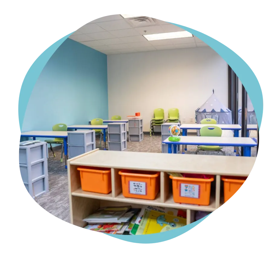 Bright classroom with green chairs, blue desks, storage bins, and a play tent in the corner.