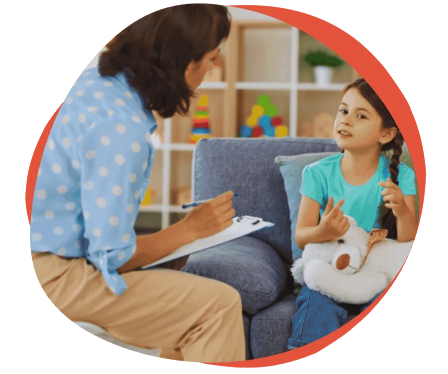 Child talking to therapist on couch while holding a teddy bear in a colorful playroom setting