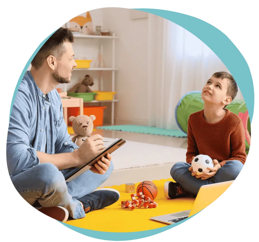 Therapist sitting with clipboard interacting with young boy holding a toy ball in a colorful playroom.