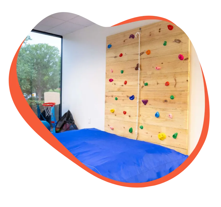 Indoor rock climbing wall with colorful handholds and blue padded floor in a bright playroom with window.